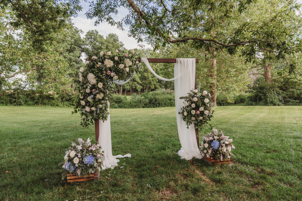 Outdoor wedding ceremony at Kennedy Estate with ivory-draped wooden arbor and purple and blue flowers